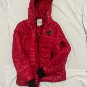 Röd Moncler pufferjacka  - Säljer en röd pufferjacka från Moncler med huva och svart logopatch på bröstet. Jackan har quiltad design, svarta muddar och dragkedja framtill. Insidan har en cool serietidningsetikett och flera detaljer. Perfekt för dig som vill ha en snygg och varm jacka.