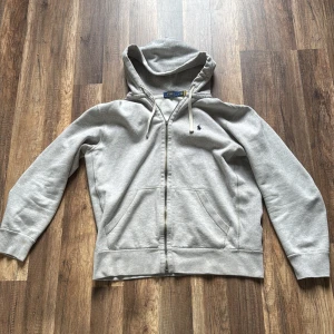 Grå hoodie från Polo Ralph Lauren - Snygg ljusgrå hoodie från Polo Ralph Lauren med dragkedja, huva med snörning och klassisk blå logga broderad på bröstet. Tillverkad i mjuk bomull och har två stora fickor framtill. Perfekt för chill dagar eller när du vill ha en clean look. Enda defekten är att den saken man drar i dragkedjan är sönder. Den är äkta.