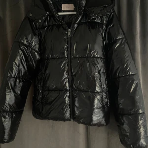 Svart pufferjacka från Nelly - Snygg svart pufferjacka från Nelly med glansig yta och huva. Jackan har dragkedja och är fodrad med mjuk fleece på insidan av huvan och i fickorna. Jag har knappt använd den så den är som ny. Kontakta mig gärna vid frågor 😊