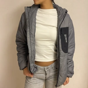 Grå pufferjacka från Polaris - Säljer en grå pufferjacka från Polaris med svart bröstficka och huva. Jackan har dragkedja framtill och quiltad design, samt en snygg kontrastdetalj på fickan med logotyp. Perfekt för dig som gillar sportig och clean stil.