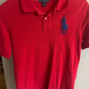 Röd pikétröja fr Polo Ralph Lauren  - Barnstorlek L som motsvarar 12 år storlek 152 cm.                                 Röd pikétröja från Ralph Lauren med klassisk krage, två knappar och stor blå broderad polospelare på bröstet. På ena ärmen finns en blå applikation med siffran 3. Tröjan är i bomull och har korta ärmar. Perfekt för en sportig och stilren look. 