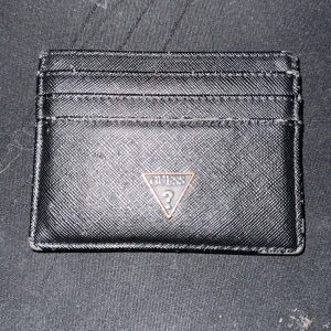 kortfodral från Guess/IT DISKUSSION - Slimmat svart kortfodral från Guess med klassisk trekantslogga i metall på framsidan. Fodralet har flera kortfack och är gjort i ett tåligt syntetmaterial med lätt textur. Perfekt för dig som vill ha koll på korten utan att ta plats i fickan.