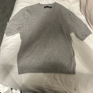 Grå stickad kortärmad tröja - Säljer en stilren grå stickad tröja från lager 157 med korta ärmar och rund hals. Perfekt för lager-på-lager och enkel att matcha med olika outfits. Tröjan har en mjuk känsla och ribbade kanter vid ärmslut och nederkant.