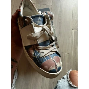 Valentino Rockstud sneakers med mönster - Unika Valentino Rockstud sneakers i skinn med coolt blommigt mönster i blått, vitt, rött och beige. Skorna har klassiska nitar baktill, vita snören och platt sula. Perfekta för dig som vill sticka ut med exklusiva detaljer och färgstark design. Strl 36 1/2
