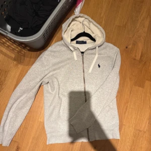 Grå hoodie från Polo Ralph Lauren - Snygg ljusgrå hoodie från Polo Ralph Lauren med dragkedja och vit snörning i huvan. Klassisk broderad logga på bröstet och mjukt innerfoder i huvan. Perfekt för chill dagar eller när du vill ha en clean look. Pris kan sänkas vid snabb affär☺️!