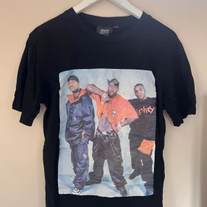 Naughty by Nature svart t-shirt - Svart t-shirt från Naughty by Nature med stor färgglad print av hiphopgrupp på framsidan och röd logga på ryggen. Klassisk passform och rund hals. Perfekt för dig som gillar streetwear och old school vibes.