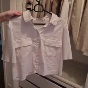 Vit kortärmad skjorta med fickor - Vit skjorta med korta ärmar och två stora bröstfickor med lock. Skjortan har klassisk krage och knappar framtill. Modellen är lite boxig och croppad, vilket ger en modern vibe. Perfekt för dig som gillar stilrena och enkla plagg.