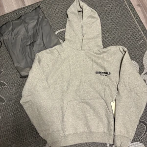 Grå Essentials Fear of God hoodie - Snygg grå hoodie från Essentials Fear of God med svart logga på bröstet och stor text på ryggen. Klassisk känguruficka framtill och stor huva. Mjuk insida och relaxed fit, perfekt för chill dagar. Kommer med originalpåse och tag kvar.