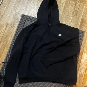 Svart oversized Nike hoodie - Svart hoodie från Nike i oversized fit med klassisk logga på bröstet. Tröjan har huva med snörning och en stor magficka. Perfekt för chill dagar och streetstyle. Mjuk och skön känsla tack vare bomullsmaterialet.