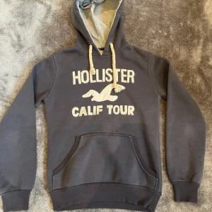 Mörkblå Hollister hoodie  - Snygg mörkblå hoodie från Hollister med broderad logga och texten 'CALIF TOUR' i vitt på bröstet. Tröjan har en stor huva med grova snören, känguruficka och ribbade muddar. Tillverkad i 100% bomull för skön känsla! Skicket är även grymt bra! Hör av vid fler frågor!