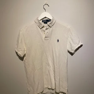 Vit pikétröja från Polo Ralph Lauren - Vit pikétröja från Polo Ralph Lauren i slim fit-modell. Klassisk krage, korta ärmar och knäppning framtill. Tröjan har den ikoniska broderade loggan på bröstet och är tillverkad i 100% bomull för en skön och fräsch känsla.