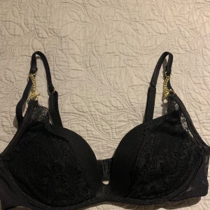 Svart spets-bh från Intimissimi 75B - Snygg svart bh från Intimissimi med spetsdetaljer och guldfärgade kedjor på axelbanden. Kuporna är vadderade och ger en fin form. Perfekt för dig som gillar en elegant och trendig look. Storlek 75B (EU), 34B (UK/USA).