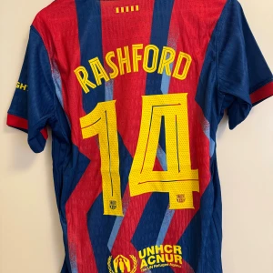 Barcelona Rashford #14 fotbollströja Nike - Snygg Barcelona fotbollströja med Rashford och nummer 14 på ryggen. Tröjan har röda och blå blixtmönster, gula detaljer och klubbmärke framtill. Tillverkad i lätt och ventilerande material, perfekt för match eller träning.