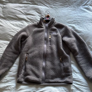 Grå fleece från Patagonia - Mysig grå teddyjacka från Patagonia med hel dragkedja framtill, hög krage och tre fickor med dragkedja. Jackan är tillverkad i fleece. Knappt använd och väldigt bra skick. 