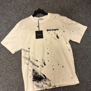 Palm Angels vit t-shirt med tryck - Säljer en vit t-shirt från Palm Angels med svart logotyptryck fram och bak. T-shirten har coola svarta färgstänk och mönster som ger en edgy vibe. Klassisk rund hals och korta ärmar. Materialet är mjuk bomull som känns skönt mot huden.