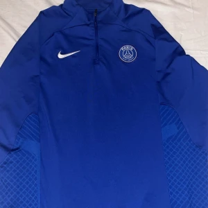 Blå Nike PSG långärmad tröja - Snygg blå långärmad tröja från Nike med Paris Saint-Germain-logga på bröstet och vit Nike-swoosh. Tröjan har half zip och sportig design med diskret mönster på sidorna. Perfekt för dig som gillar fotboll och vill ha en clean look.