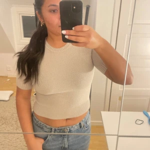 Beige stickad croppad topp från Zara - Säljer en beige stickad croppad topp från Zara med korta ärmar och rund hals. Toppen har en ribbad kant vid halsen, ärmslut och nederkant samt ett diskret stickat mönster över hela plagget. Perfekt att styla med jeans eller kjol för en chill look.