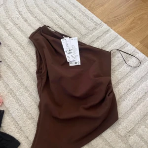 Brun one shoulder klänning XS - Säljer en snygg brun one shoulder topp i storlek XS. Toppen har en asymmetrisk axel med smal axelrem och är tillverkad i ett mjukt, stretchigt material som sitter tight och markerar figuren.
