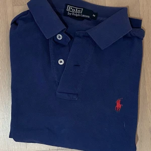 Marinblå pikétröja från Polo Ralph Lauren - Snygg marinblå pikétröja från Polo Ralph Lauren med klassisk krage och två vita knappar framtill. Tröjan har korta ärmar och den ikoniska röda Polo-loggan broderad på bröstet. Tillverkad i mjuk bomull som är skön mot huden.