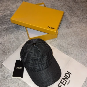 Svart Fendi keps med FF-mönster - Snygg svart keps från Fendi med ikoniskt FF-mönster över hela kepsen. Justerbar rem baktill i svart läder med Fendi-logga. Kepsen är tillverkad i bomull och har en klassisk böjd skärm. Perfekt för dig som vill ha en lyxig och trendig accessoar.