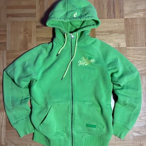 Grön hoodie från Peak Performance - Snygg grön hoodie från Peak Performance med dragkedja, huva och snörning. Broderad logga på bröstet och liten logga på huvan. Mjuk insida och känguruficka framtill. Perfekt för chill eller träning.