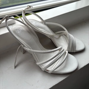 Vita sandaler med klack från Zara - Säljer ett par stilrena vita sandaler från Zara med smala remmar och öppen tå. Skorna har en elegant klack och spänne runt ankeln. Perfekta för dig som gillar minimalistisk och trendig design. Materialet är syntet och de är riktigt lätta att matcha. Perfekt till studenten!