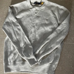 Grå sweatshirt från Polo Ralph Lauren - Snygg ljusgrå sweatshirt från Polo Ralph Lauren med klassisk rund halsringning och diskret broderad logga på bröstet. Tröjan är i mjuk bomull och har ribbade muddar vid ärmslut och nederkant. Perfekt för en clean och stilren look.