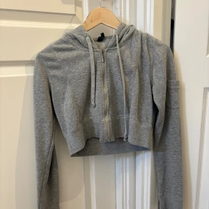 Grå croppad hoodie med dragkedja - Grå croppad hoodie med dragkedja framtill och snörning i huvan. Långa ärmar och liten ficka på ena ärmen.💕