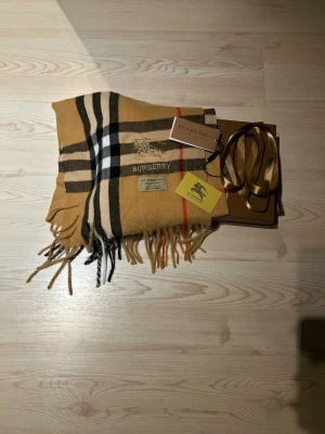 Halsduk från Burberry - Exklusiv halsduk från Burberry i klassiskt rutig design med beige, svart, vit och röd färg. Halsduken har broderad Burberry-logga framtill. Snygga fransar nedtill och medföljande originalförpackning och tags.