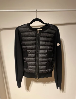 Svart cardigan jacka från Moncler - Svart cardigan jacka från Moncler med quiltad front i syntetmaterial och stickade ärmar samt rygg. Rund hals, dragkedja framtill och Moncler-logga på vänster ärm. Perfekt för dig som gillar stilren design och exklusiva detaljer. Cardigan i gott skick, låda medföljer.