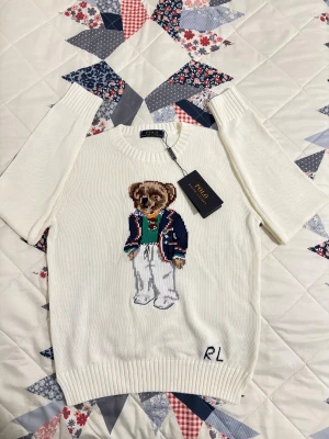 Vit stickad tröja Polo Ralph Lauren - Vit stickad tröja från Polo Ralph Lauren med klassisk rund hals och ribbade muddar. Framsidan pryds av en broderad nalle i preppy outfit och RL-broderi nere vid kanten. Perfekt för dig som gillar ikoniska detaljer och stilren design. Storleken är L men passar som S. Priset kan diskuteras 🥂