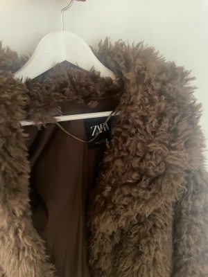 Brun teddyjacka från Zara - Mjuk och fluffig brun teddyjacka från Zara med krage och helfoder. Jackan har en oversized look och är perfekt för att hålla sig varm under kyliga dagar. Materialet är pälsimitation som ger en cool och trendig vibe.