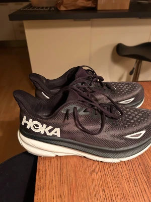 Hoka Clifton 9 svarta sneakers 46 - Säljer ett par Hoka Clifton 9 sneakers i svart med vita detaljer och logga på sidan. Skorna har snörning, rund tå och en tjock, vit platt sula som ger grym komfort. Ovandelen är i syntetmaterial med mönstrad mesh för bra ventilation. Perfekta för träning och löpning.