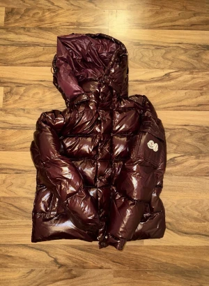Mörk vinröd pufferjacka från Moncler - Säljer en mörk vinröd pufferjacka från Moncler med glansig finish och huva. Jackan har quiltad design, Moncler-logga på ärmen och dragkedja framtill. Perfekt för kalla dagar och har en riktigt schysst streetstil.