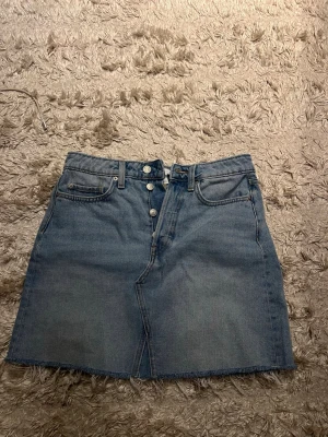 Blå jeanskjol från H&M, strl 36 - Säljer en blå jeanskjol från H&M i storlek 36. Kjolen har klassisk femficksdesign, knappar framtill och rå kant nedtill för en cool look. Perfekt att matcha med sneakers eller boots. Materialet är bomull och kjolen har en normal passform.