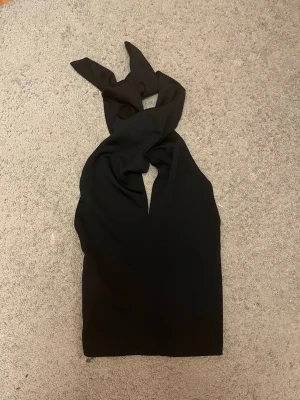 Svart topp med knyt - Säljer en svart topp med omlott och knyt i halsen. Toppen är ärmlös och har en croppad passform med elastisk rygg för extra komfort. Perfekt för dig som gillar stilrena och trendiga plagg med snygga detaljer.