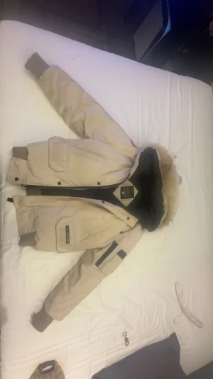 Beige Canada Goose parkajacka med päls - Säljer en beige Canada Goose parkajacka med äkta päls på huvan och snygga detaljer. Jackan har flera fickor, ribbade muddar och dragkedja med knappar. Perfekt för kalla dagar och har den klassiska Canada Goose-loggan på ärmen. Passar 175com