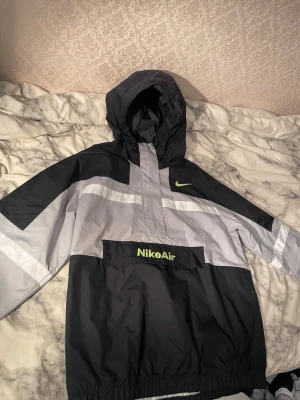 Nike Air vindjacka svart/grå M - Nike Air vindjacka i svart och grått med stora loggor i limegrönt på bröstet och fickan. Jackan har huva, elastisk nederkant och en stor ficka framtill. Materialet är polyester och jackan har en sportig, loose passform. Perfekt för dig som gillar streetwear.