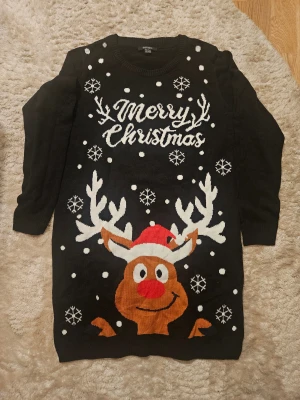 Svart jultröja med renmotiv från Esmara - Rolig svart stickad jultröja från Esmara i storlek M. Framsidan har ett stort motiv med en ren i tomteluva, snöflingor och texten 'Merry Christmas' i vitt. Tröjan har rund hals och långa ärmar, perfekt för julmys och vinterhäng.