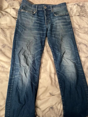 G-Star RAW 3301 Straight jeans blå - Säljer ett par G-Star RAW 3301 Straight jeans i klassisk blå tvätt med snygga slitningar och kontrastsömmar. Modellen har raka ben, fem fickor och detaljerad logopatch i brunt läder bak. Perfekt för dig som gillar street och enkel stil.