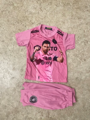 Rosa Inter Miami Messi fotbollströja - Säljer ett rosa fotbollskit med tröja och shorts från Inter Miami. Tröjan har tryck med spelare på framsidan, svarta detaljer och Messi 10 på ryggen. Shorts med klubbmärke och resår i midjan. Perfekt för unga fotbollsfans!