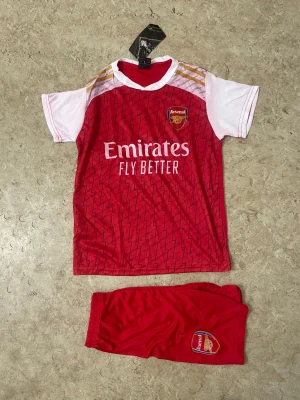 Arsenal fotbollströja G.Jesus #9 - Säljer en röd och vit Arsenal fotbollströja med G.Jesus och nummer 9 på ryggen. Framsidan har Emirates Fly Better-tryck och klubbmärke. Tillhörande röda shorts med Arsenal-logga ingår. Tröjan har korta ärmar och mönstrad framsida.