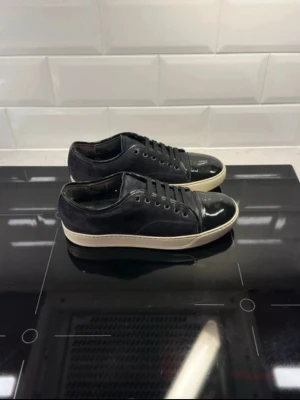 Svarta Lanvins skor 41 - Säljer ett par svarta sneakers med snygg lackad tå och vit platt sula. Skorna har snörning och är gjorda i mocka med detaljer i syntet. Perfekt för dig som gillar stilrena och coola skor med en twist.