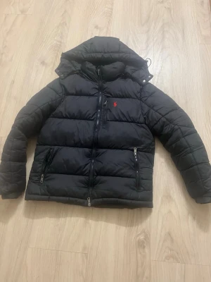 Svart pufferjacka från Polo Ralph Lauren - Svart pufferjacka från Polo Ralph Lauren med huva, dragkedjor på fickorna och bröstet samt det klassiska röda Polo-logot på vänster sida. Jackan är quiltad och har långärmade armar, perfekt för kalla dagar. Materialet är polyester och jackan har en loose passform. Priset kan diskuteras 
