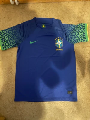 Blå Brasilien fotbollströja Nike - Snygg blå Brasilien fotbollströja från Nike med grönt Nike-märke och Brasilien-emblem på bröstet. Ärmarna har leopardmönster i grönt och blått med gula detaljer vid mudden. Perfekt för dig som älskar fotboll och vill sticka ut med cool design.