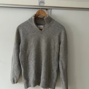 Grå merinoulltröja med half zip från Vailent - Snygg grå stickad tröja från Vailent i mjuk merinoull. Tröjan har en ribbad hög krage och en praktisk half zip framtill. Långärmad och med normal passform, perfekt för dig som gillar stilren och enkel design. Tröjan är använd ett fåtal gånger. Tröjan är i storlek small men jag skulle säga att den är i M då den passar mig bra!