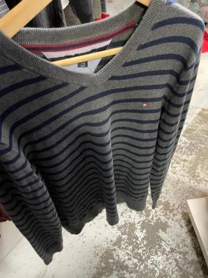 Randig grå tröja från Tommy Hilfiger - Snygg grå tröja från Tommy Hilfiger med mörkblå ränder och diskret logga på bröstet. Tröjan har v-ringad hals och ribbade muddar vid ärmslut och nederkant. Perfekt för dig som gillar klassisk stil med en modern touch.