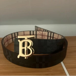  Burberry bälte  - Snyggt svart bälte från Burberry med klassiskt rutigt mönster och ett stort guldigt B-spänne framtill. Insidan har Burberrys signaturmönster i beige och rött. Perfekt för att lyfta din outfit med en lyxig touch.