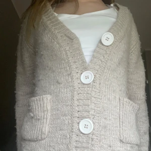 Beige stickad kofta från Gul&Blå - Mysig beige stickad kofta från Gul&Blå med stora vita knappar och två fickor framtill. Koftan har lång ärm och en klassisk v-ringad design, perfekt att slänga på sig över en t-shirt eller topp.