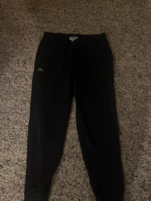 Svarta mjukisbyxor från Lacoste Sport - Svarta mjukisbyxor från Lacoste Sport med klassisk krokodillogga på benet. Byxorna har elastisk midja och muddar vid bensluten.  tillverkade i mjukt bomullsmaterial.